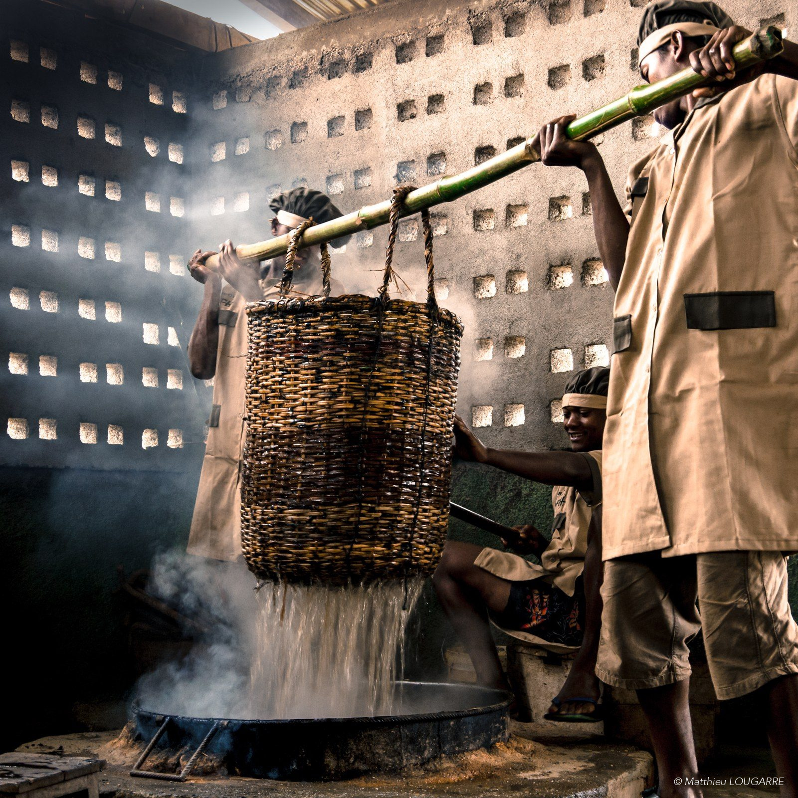Three workers lift a wicker basket of vanilla beans out of a steaming hot-water tank during killing, the first step of curing.